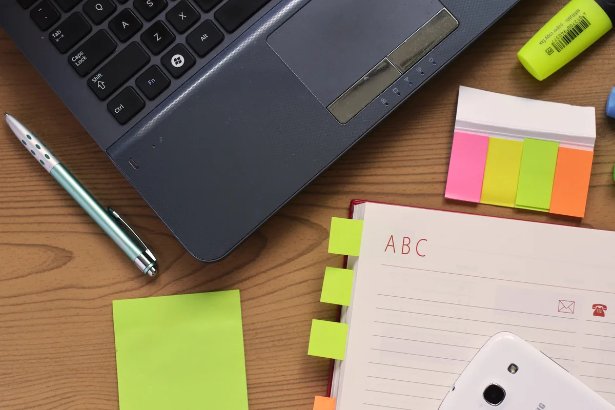 Laptop keyboard, pen, colorful page tabs and an open notebook on a wooden desk.