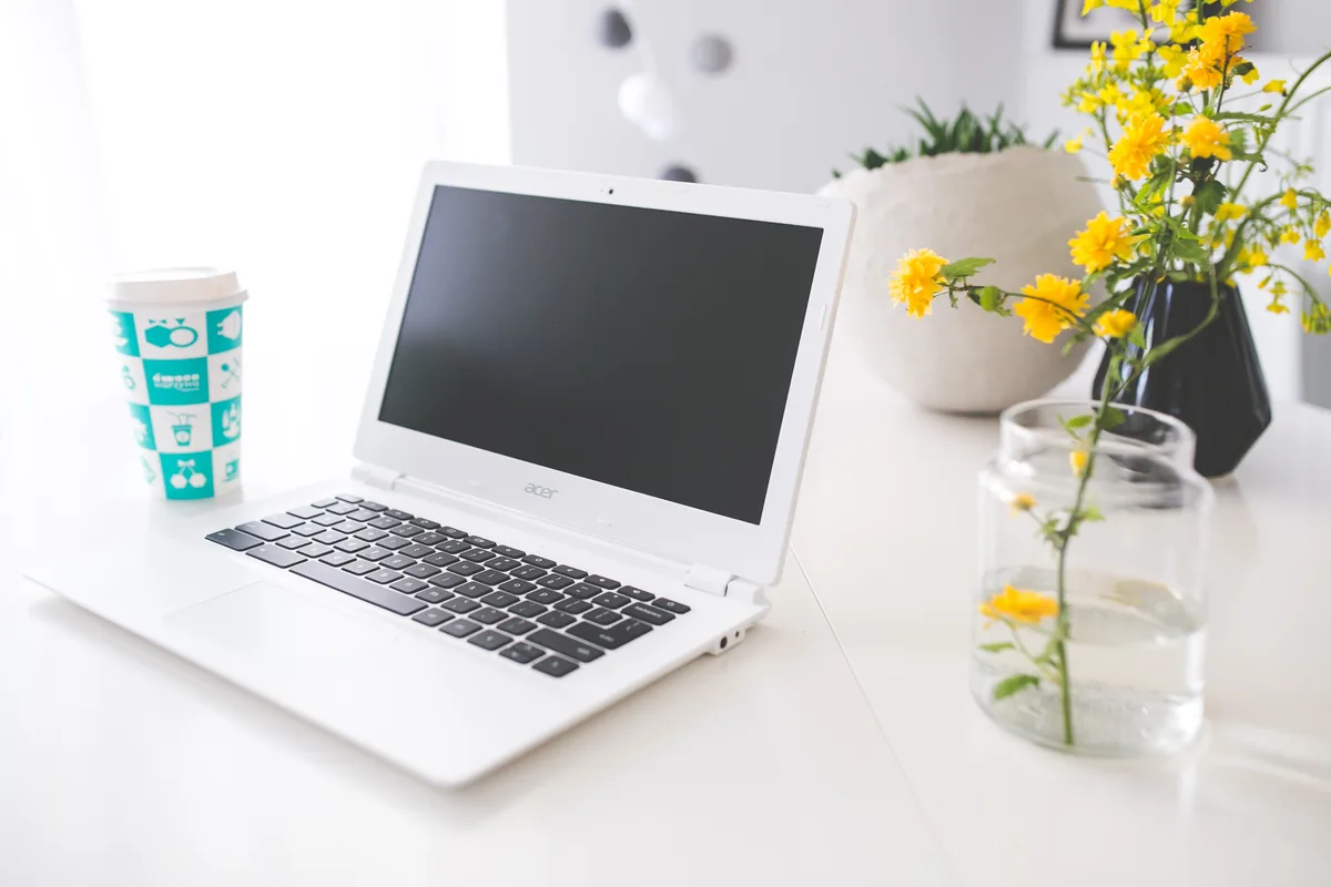 White laptop with a blank screen, a takeaway cup and yellow flowers on a white desk.